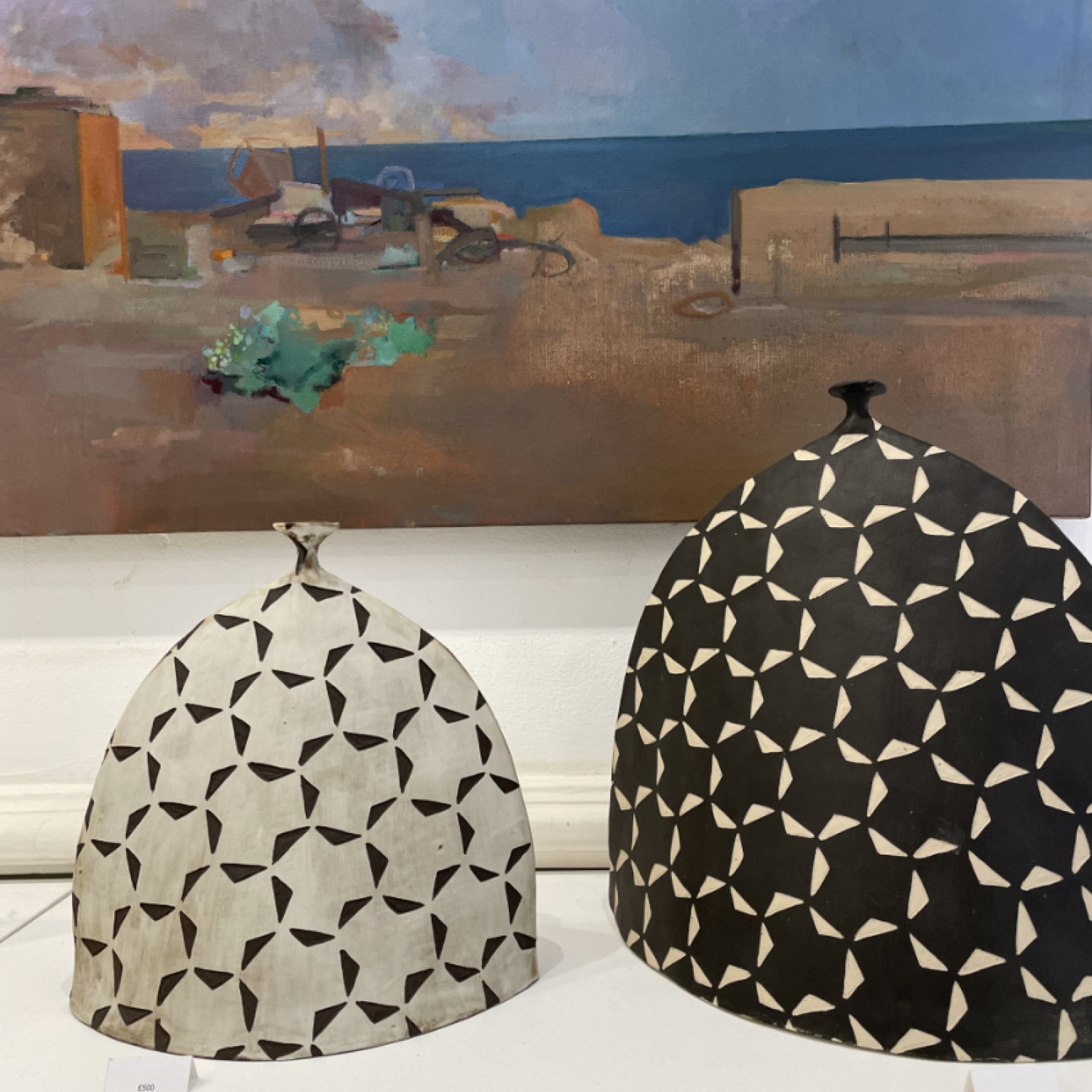 Two black and white handmade ceramic vessels. Behind a section of a seascape painting with blue sky and clouds.