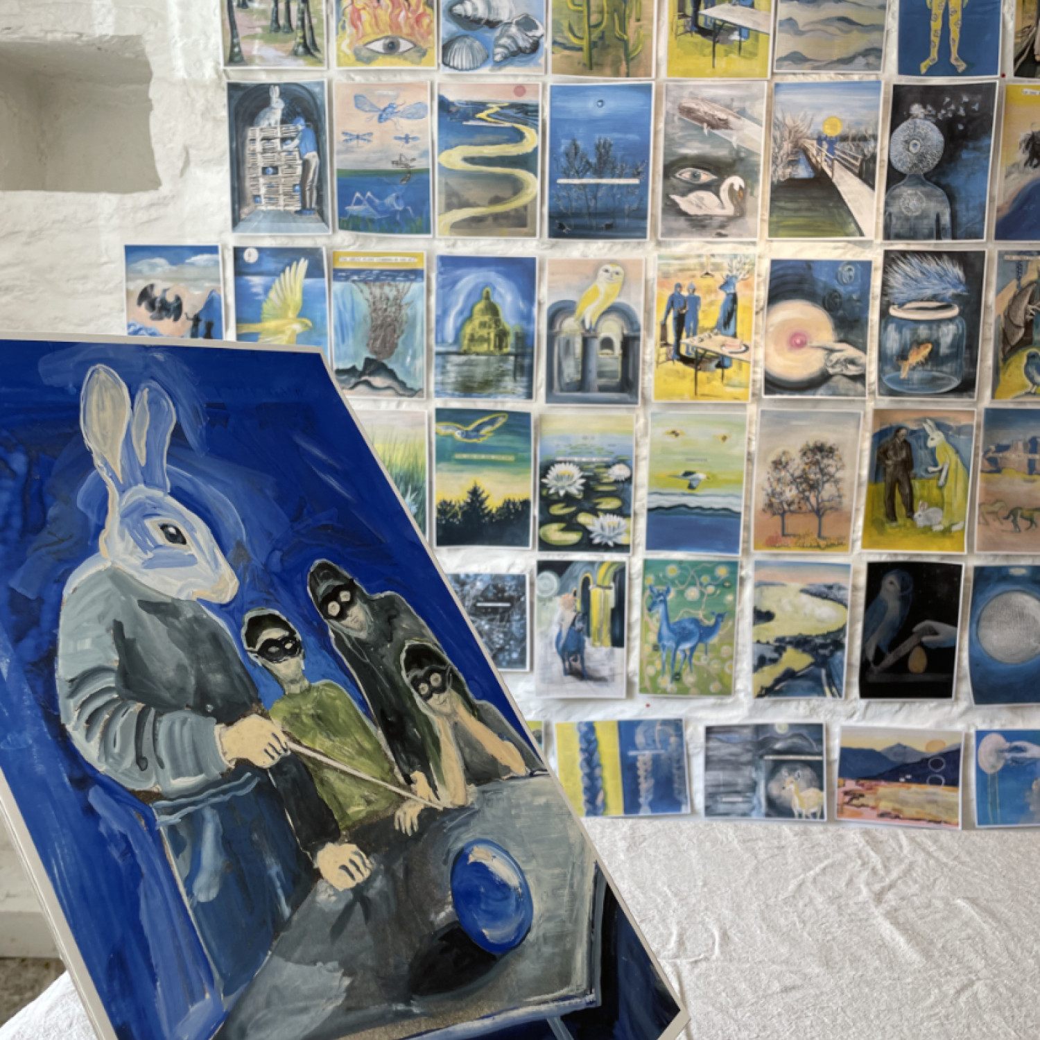 Foreground, left, a painting of a person with a rabbits head holding a wand, pointing at a blue orb on a grey table. On the right, three young people wearing black eye masks. Background shows a wall covered in various images, mainly in tones of yellow and blue.
